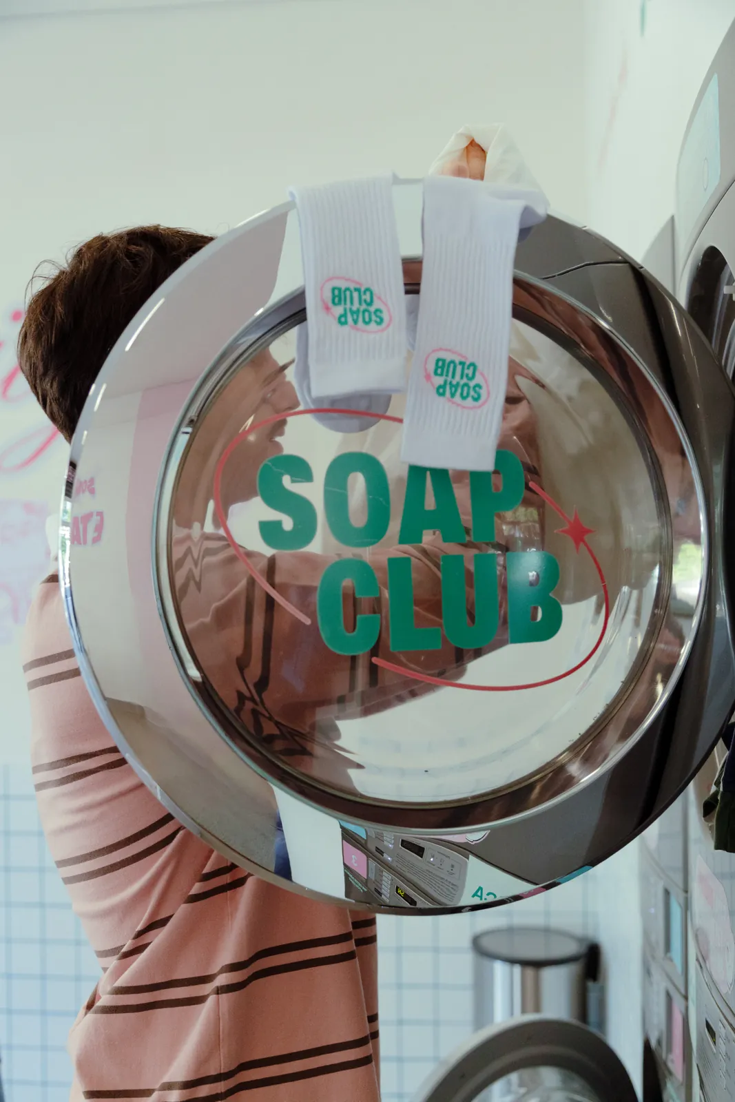 Pessoa atrás da porta aberta de uma lavadora com o logo SOAP CLUB em verde e duas meias brancas da marca penduradas