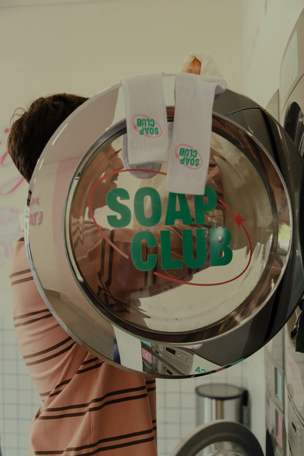 Pessoa pendura meias brancas do SOAP CLUB na porta cromada de uma máquina de lavar com o logo "SOAP CLUB" em verde