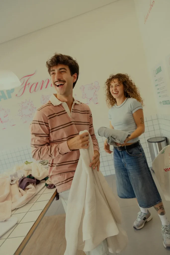 Homem e mulher rindo enquanto dobram roupas na bancada do SOAP CLUB, com mural "Soap Fam" e mascotes ao fundo