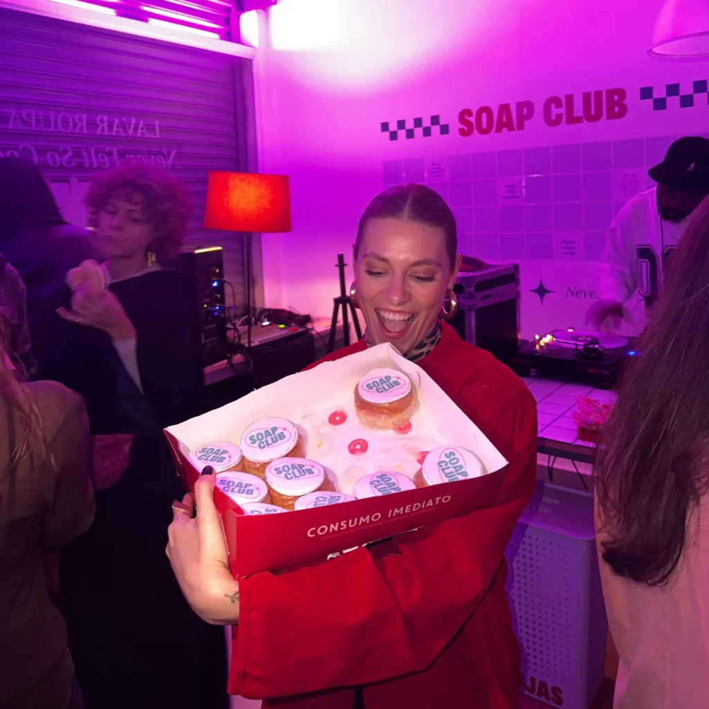 Mulher sorrindo segura caixa de donuts com logo SOAP CLUB em festa na loja, luz rosa, DJ ao fundo